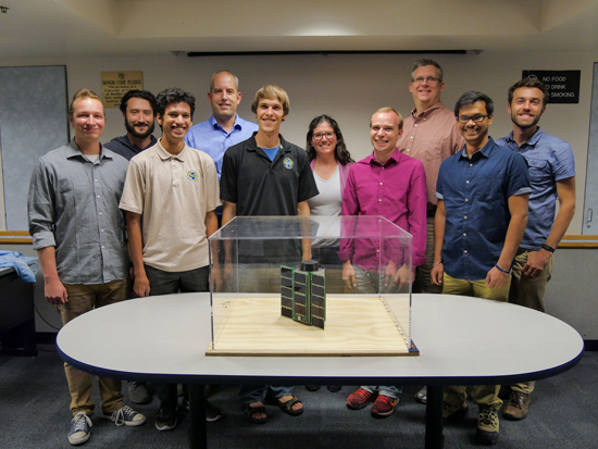 Members of team stand behind the QB-50 student-built microsatellite cube. The satellite is headed to the International Space Station as part of an international effort to study the Earth’s atmosphere.