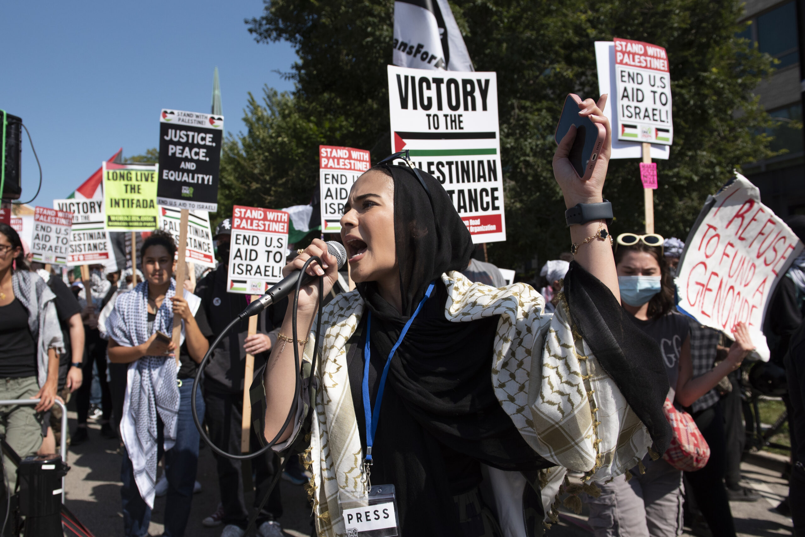 A young woman wearing a hijab and a keffiyeh speaks into a microphone to pro-Palestine protestors outside of the DNC. Protest signs read: End U.S. Aid to Israel.