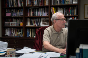 A man sits with his hands typing on a computer in an academic office, a full bookshelf behind him he has short cut grey hair and black glasses, wearing a casual polo.