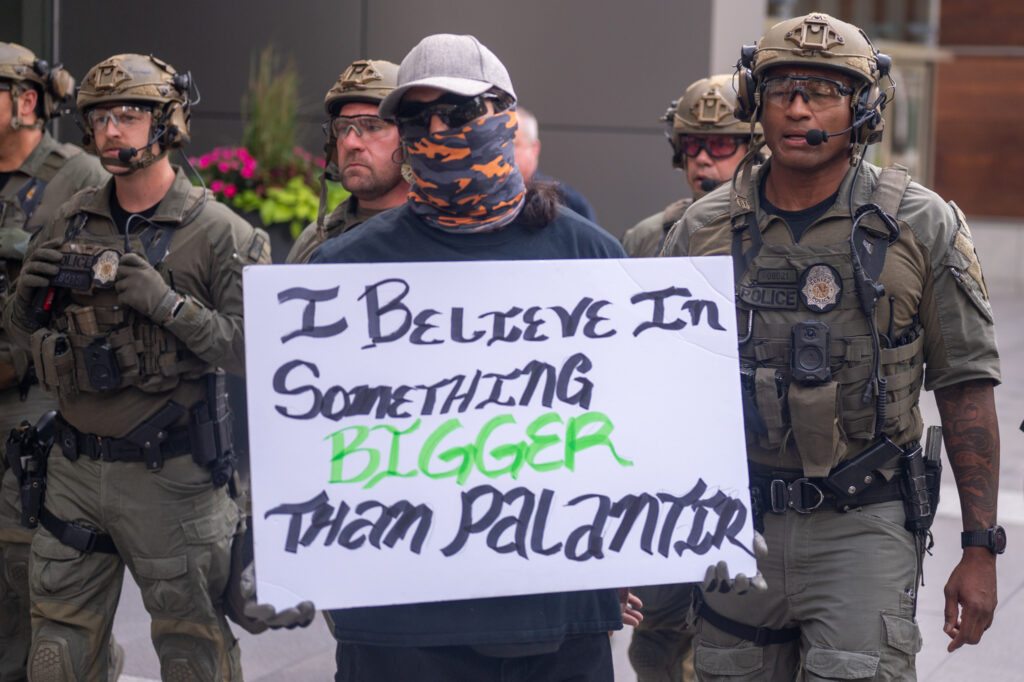A masked protestor carries a sign reading "I believe in something bigger than Palantir" in front of several police officers wearing military-style camouflage clothing.