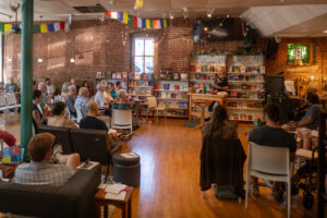 In a warmly lit room, below Tibetan prayer flags, a coffee shop and book store space has been converted temporarily in to a worship space, tables and chairs all pointed to the center of the room where a woman with white hair speaks.