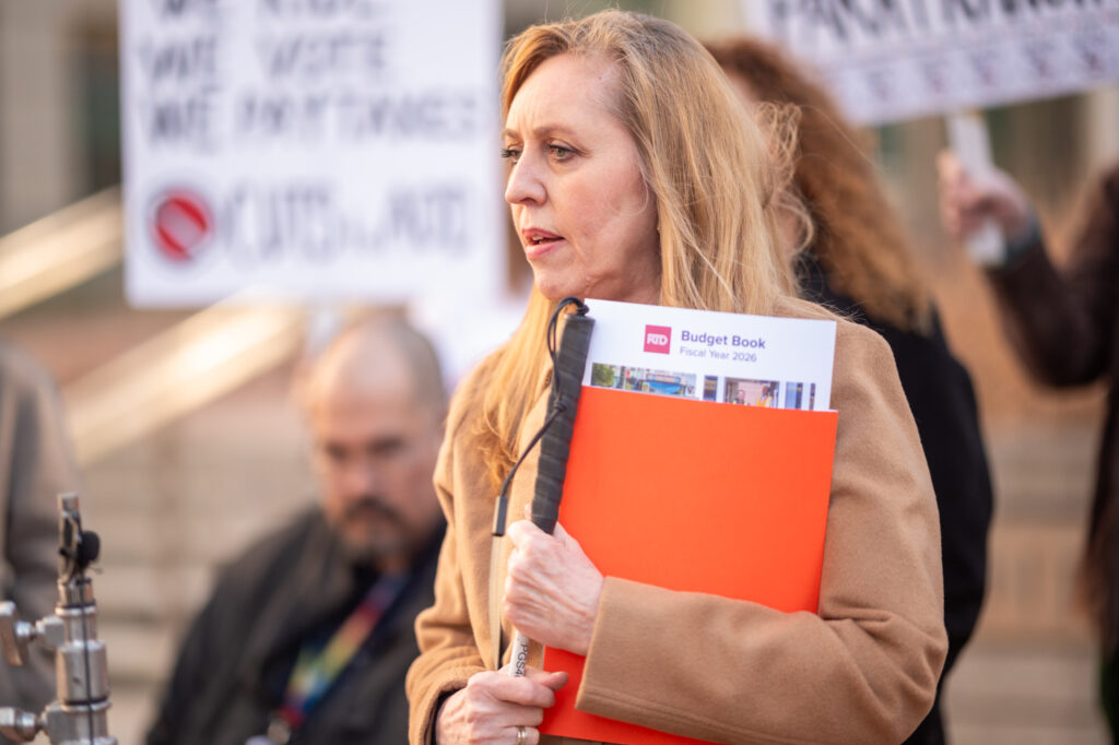 Standing and holding an orange folder with the RTD 2026 Budget Book poking above the top and her mobility cane, a blonde woman in a brown jacket speaks before a crowd holding protest signs. 