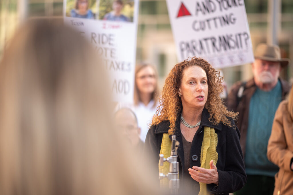With a serious look on her face, projecting that she is making a point, a woman with curly red hair speaks before a crowd holding protest signs during a press conference. 