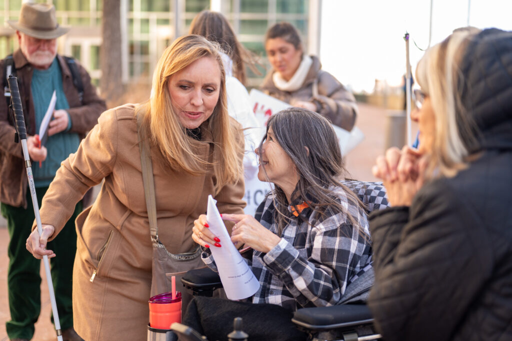 Wearing a black and white plaid shirt, a woman with long grey hair speaks from her wheelchair to a woman leaning forward to hear, her hair is chest length and blonde, wearing a knee length brown jacket. 